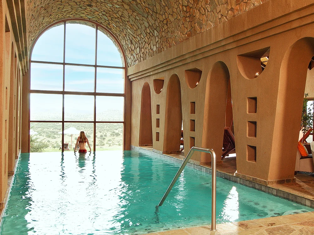The heated indoor swimming pool in the wellness area of GocheGanas Lodge, featuring a large arched window offering views of the outdoor landscape. The pool area has a high, stone-accented ceiling and arched alcoves along the side. A person in a red swimsuit is partially visible in the water near the window.